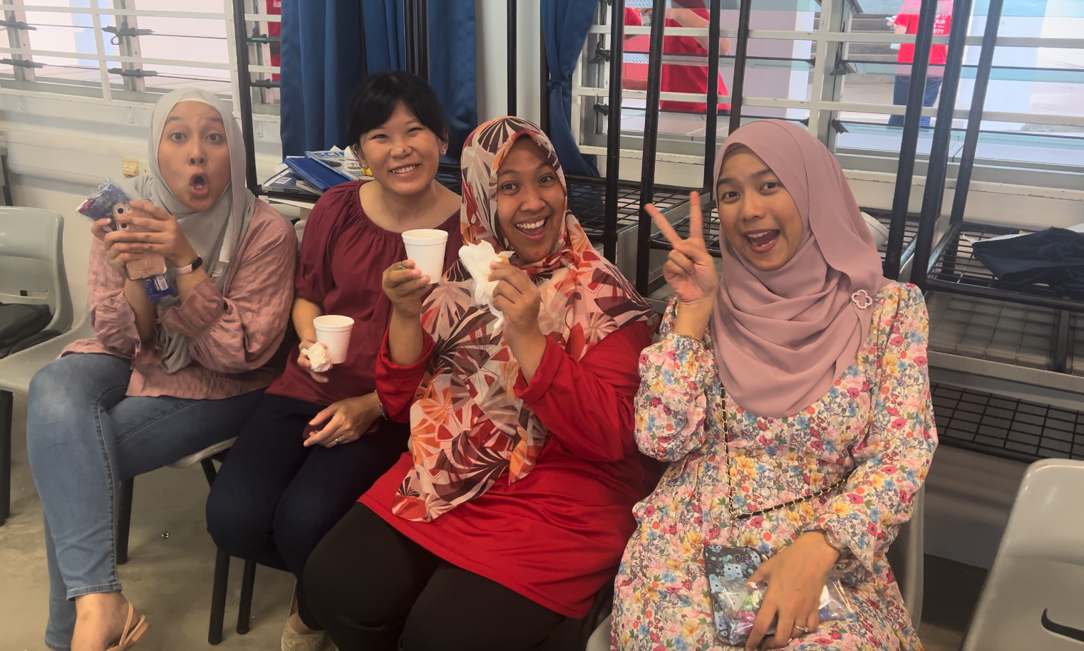 Four smiling women sit together indoors, three wearing hijabs, holding cups and snacks, one making a peace sign.